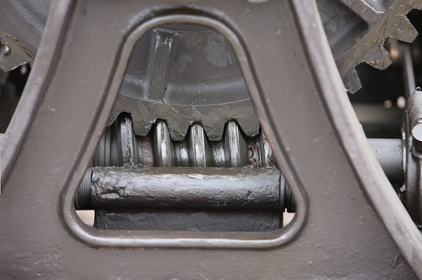 Close-up view of a mechanical gear system, featuring large metal components with visible gears and axles. The metal surfaces are painted in a matte gray color, with signs of wear and some accumulated grease at the joints.