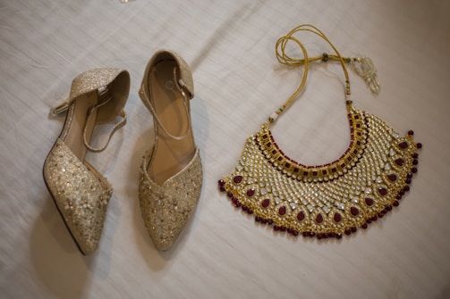A pair of sparkling, gold-colored heels adorned with glitter and embellishments next to a richly designed necklace on a white textured surface. The necklace features intricate patterns with red and gold accents, showcasing a luxurious and traditional style.