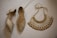A pair of sparkling, gold-colored heels adorned with glitter and embellishments next to a richly designed necklace on a white textured surface. The necklace features intricate patterns with red and gold accents, showcasing a luxurious and traditional style.