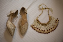 A pair of sparkling, gold-colored heels adorned with glitter and embellishments next to a richly designed necklace on a white textured surface. The necklace features intricate patterns with red and gold accents, showcasing a luxurious and traditional style.