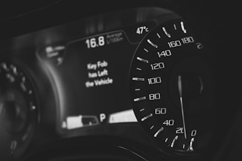 A close-up of a car dashboard displays a speedometer with numbers ranging from 0 to 220. The digital screen shows a temperature of 47°C and a message indicating that the key fob has left the vehicle. The dashboard features sleek, modern design elements with monochrome colors.