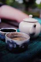 A ceramic teapot and two cups are placed on a green textured surface. The teapot is off-white with a simple, smooth design, while the cups have a rustic, earthy appearance with a mix of blue, gray, and beige tones. The background is slightly blurred with hints of a natural, outdoor setting.