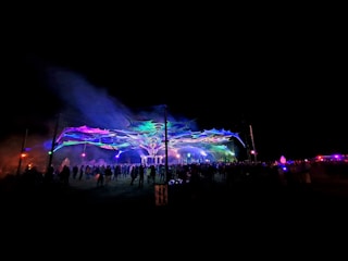 A vibrant outdoor nighttime scene featuring a large crowd gathered under a colorful, illuminated canopy. The canopy has psychedelic patterns and is highlighted by various neon lights, creating a festive atmosphere.