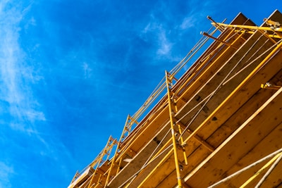 A construction scaffold rises up against a clear blue sky. The yellow metal framework is vertically oriented, creating a sense of height and structure. The wooden platforms are visible between the metal supports.
