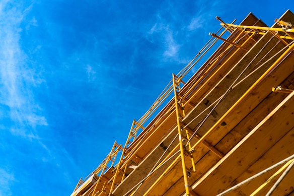 A construction scaffold rises up against a clear blue sky. The yellow metal framework is vertically oriented, creating a sense of height and structure. The wooden platforms are visible between the metal supports.
