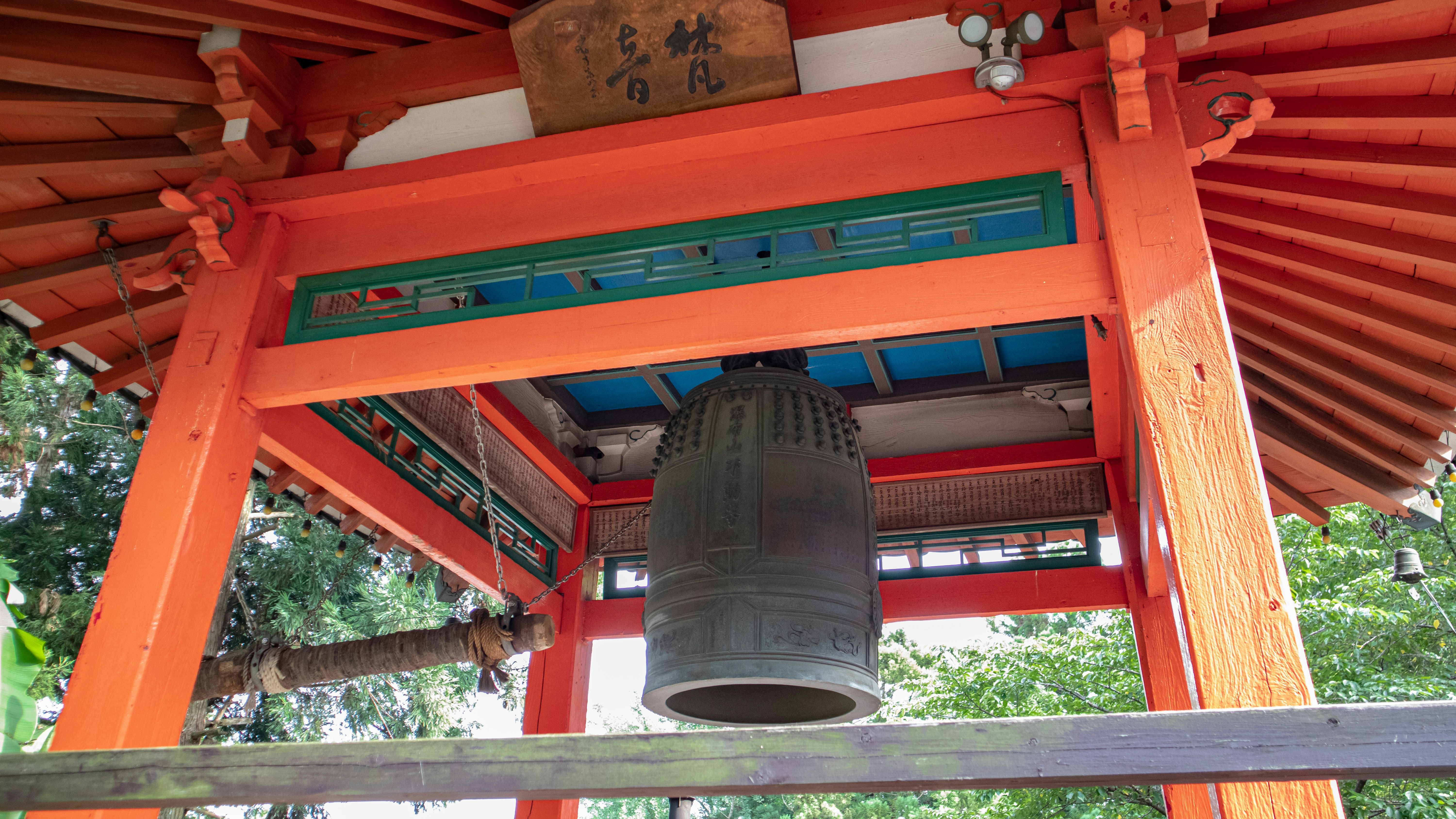 Large bronze bell hangs centered inside a vivid red timber pavilion, framed by blue-green interior beams and carved signage above.