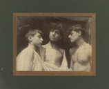 Three young men are gathered closely together, each with neutral expressions. They are topless, with light fabric draped over their shoulders. The background is dark and textured, creating a stark contrast with their skin tones.