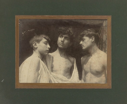 Three young men are gathered closely together, each with neutral expressions. They are topless, with light fabric draped over their shoulders. The background is dark and textured, creating a stark contrast with their skin tones.