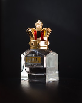 A glass bottle of perfume with a luxurious, ornate golden crown-shaped cap. The label on the bottle reads 'SCANDAL' in capital letters with a gold and black color scheme.