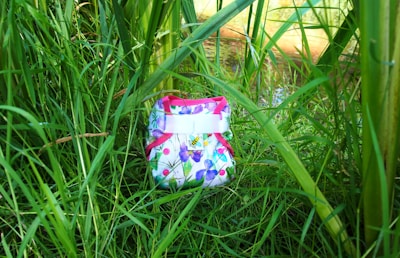 A colorful cloth diaper with a floral pattern is nestled among tall green grass and plants. The diaper features pink trim and buttons with a bright, nature-inspired design.