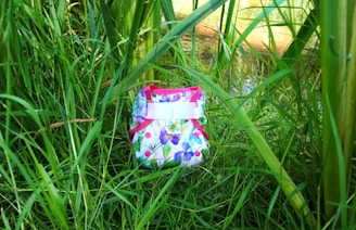 A colorful cloth diaper with a floral pattern is nestled among tall green grass and plants. The diaper features pink trim and buttons with a bright, nature-inspired design.