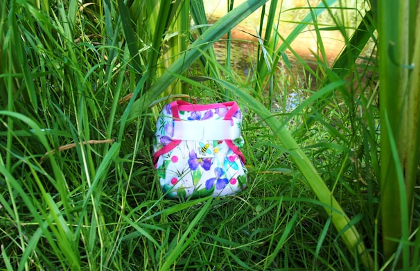 A colorful cloth diaper with a floral pattern is nestled among tall green grass and plants. The diaper features pink trim and buttons with a bright, nature-inspired design.