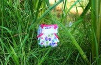 A colorful cloth diaper with a floral pattern is nestled among tall green grass and plants. The diaper features pink trim and buttons with a bright, nature-inspired design.