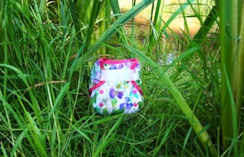 A colorful cloth diaper with a floral pattern is nestled among tall green grass and plants. The diaper features pink trim and buttons with a bright, nature-inspired design.