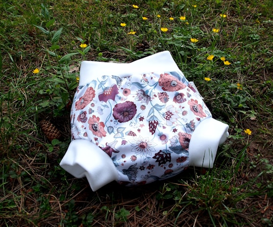 A cloth diaper with a floral pattern lies on the grass surrounded by small yellow flowers and green foliage. The diaper features a design of various flowers in pink, purple, and blue tones, framed by a white border.