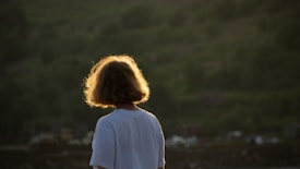 A person with short curly hair is wearing a white shirt and standing outdoors, facing away from the camera. The scene is backlit by the sun, which creates a glowing halo around the person's head. The background consists of blurred greenery, suggesting a natural setting.