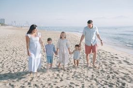 A family of five is walking on a sandy beach near the ocean. The father and mother are holding hands with their three children. The mother is wearing a long light blue dress, the father is wearing a light blue shirt and red shorts, and the children are dressed in light-colored clothing. The background shows the shoreline with gentle waves and a hazy city skyline in the distance.