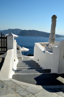 A serene view of a white staircase leading down towards a calm, blue sea. The foreground features white architecture typical of Greek islands, an umbrella, and a railing. In the background, an island is visible under a clear sky.