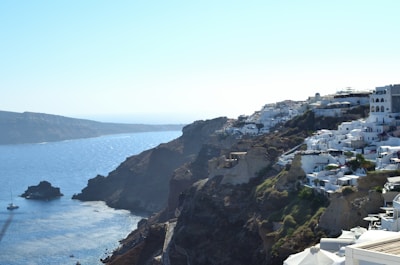 A scenic coastal view of a cluster of white buildings perched on a cliffside, overlooking a vivid blue sea. The white structures contrast beautifully with the rugged terrain, offering a classic Mediterranean aesthetic. A lone sailboat is visible in the water, adding to the tranquil coastal setting.