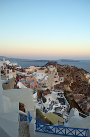 A picturesque Mediterranean village with whitewashed buildings and blue accents perched on a cliff overlooking the sea. The sky features a soft gradient from light blue to orange near the horizon. People are gathered along the pathways and terraces, possibly enjoying a sunset view.