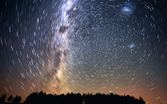 A long exposure photograph capturing the swirling motion of stars in the night sky, creating circular trails around the North celestial pole. The Milky Way is visible as a bright, nebulous band stretching across the sky. The silhouetted horizon features a dark outline of trees, contrasting with the illuminated starry sky. The lower part of the image is tinged with a subtle orange hue, suggesting the remnants of a sunset or the glow of distant city lights.