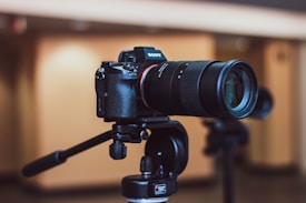 A high-end camera with a telephoto lens is mounted on a sturdy tripod, set against a softly blurred indoor background. The camera body appears robust and professional, with various buttons and dials visible. The lens features a textured grip and markings indicating focal length.