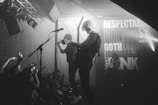 A black and white photograph captures a live band performance on stage. The scene includes musicians playing guitars and singing into microphones, with stage lights and a backdrop featuring large text like 'INDIE,' 'ROCK,' and 'PUNK.' The audience is visible in the foreground, some holding up phones capturing the moment.