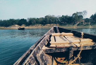 A wooden boat floats on a calm river with a view of lush green trees lining the distant riverbank. The foreground shows the boat's seating area, partially covered by wooden planks. Another boat can be seen anchored near the shore.