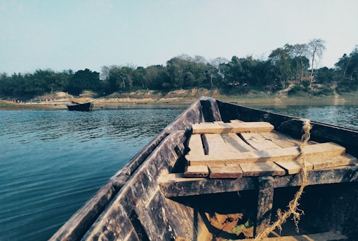 A wooden boat floats on a calm river with a view of lush green trees lining the distant riverbank. The foreground shows the boat's seating area, partially covered by wooden planks. Another boat can be seen anchored near the shore.