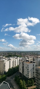 A cityscape view showcases several modern apartment buildings under a bright blue sky with fluffy white clouds. The foreground features streets lined with lush green trees, while the background extends into a vast expanse of residential areas and distant hills.