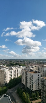 A cityscape view showcases several modern apartment buildings under a bright blue sky with fluffy white clouds. The foreground features streets lined with lush green trees, while the background extends into a vast expanse of residential areas and distant hills.