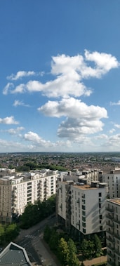 A cityscape view showcases several modern apartment buildings under a bright blue sky with fluffy white clouds. The foreground features streets lined with lush green trees, while the background extends into a vast expanse of residential areas and distant hills.
