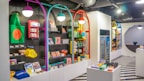 A brightly lit retail store interior featuring colorful and neatly organized merchandise displays. The walls have arches with vibrant colors like purple, teal, red, and yellow. Shelves contain a variety of items including bags, books, and small accessories. The flooring has a striped black and white pattern, and modern pendant lights hang from the ceiling.