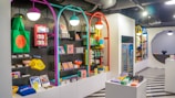 A brightly lit retail store interior featuring colorful and neatly organized merchandise displays. The walls have arches with vibrant colors like purple, teal, red, and yellow. Shelves contain a variety of items including bags, books, and small accessories. The flooring has a striped black and white pattern, and modern pendant lights hang from the ceiling.