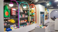 A brightly lit retail store interior featuring colorful and neatly organized merchandise displays. The walls have arches with vibrant colors like purple, teal, red, and yellow. Shelves contain a variety of items including bags, books, and small accessories. The flooring has a striped black and white pattern, and modern pendant lights hang from the ceiling.