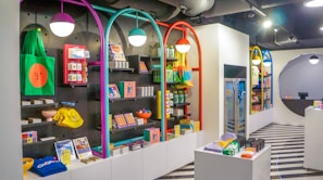 A brightly lit retail store interior featuring colorful and neatly organized merchandise displays. The walls have arches with vibrant colors like purple, teal, red, and yellow. Shelves contain a variety of items including bags, books, and small accessories. The flooring has a striped black and white pattern, and modern pendant lights hang from the ceiling.
