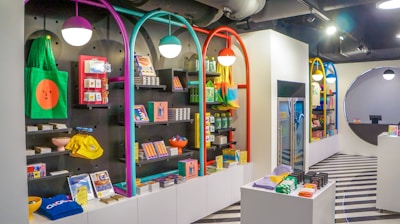 A brightly lit retail store interior featuring colorful and neatly organized merchandise displays. The walls have arches with vibrant colors like purple, teal, red, and yellow. Shelves contain a variety of items including bags, books, and small accessories. The flooring has a striped black and white pattern, and modern pendant lights hang from the ceiling.