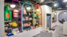 A brightly lit retail store interior featuring colorful and neatly organized merchandise displays. The walls have arches with vibrant colors like purple, teal, red, and yellow. Shelves contain a variety of items including bags, books, and small accessories. The flooring has a striped black and white pattern, and modern pendant lights hang from the ceiling.
