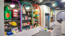 A brightly lit retail store interior featuring colorful and neatly organized merchandise displays. The walls have arches with vibrant colors like purple, teal, red, and yellow. Shelves contain a variety of items including bags, books, and small accessories. The flooring has a striped black and white pattern, and modern pendant lights hang from the ceiling.