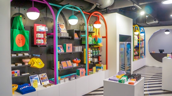 A brightly lit retail store interior featuring colorful and neatly organized merchandise displays. The walls have arches with vibrant colors like purple, teal, red, and yellow. Shelves contain a variety of items including bags, books, and small accessories. The flooring has a striped black and white pattern, and modern pendant lights hang from the ceiling.