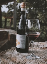 A bottle of Aglianico del Taburno wine stands on a rustic wooden surface, accompanied by a glass half-filled with red wine. The background features blurred greenery from what appears to be an outdoor setting, creating a serene atmosphere.