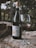 A bottle of Aglianico del Taburno wine stands on a rustic wooden surface, accompanied by a glass half-filled with red wine. The background features blurred greenery from what appears to be an outdoor setting, creating a serene atmosphere.