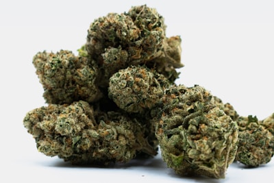 Dried cannabis buds with visible trichomes and orange pistils. The buds are dense and covered in a mixture of green and brownish colors, set against a plain white background.