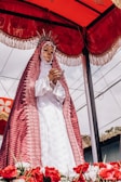 A religious statue is dressed in a richly detailed white gown with a red and intricate lace cloak. The statue wears a halo with radiating spikes and is positioned under a red canopy adorned with golden fringes. The base is decorated with vibrant red and white flowers. Power lines and a partial view of a building appear in the background, under a cloudy sky.