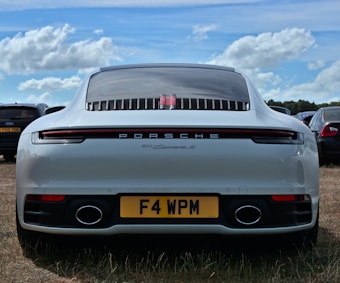 A white Porsche 911 Carrera S is parked on a grass field. The vehicle's rear features distinctive tail lights, a sleek design, and a visible license plate with the registration F4 WPM. The sky above is bright with scattered clouds, and other parked cars are visible in the background.