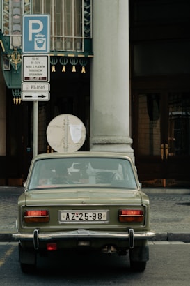 An olive green vintage car is parked on a city street, facing a decorative building facade. The car's license plate reads AZ-25-98. Next to it is a parking sign with multiple language instructions.