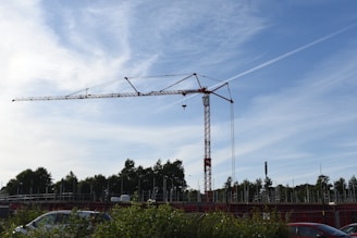 A large red crane towers over a construction site, surrounded by trees and partially obscured by bushes and vehicles. The structure is set against a backdrop of a clear blue sky with wispy clouds.