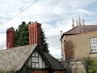 A scene featuring historic architecture with prominent red brick chimneys and slate roofs. In the foreground, a building labeled 'The Lodge 1831' is partially visible with a triangular roof section. A large evergreen tree stands beside it. To the right, another building is visible with bare vines climbing its wall. The upper part of a church with spires is seen in the background against a cloudy sky.