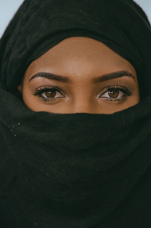 A close-up of a person with only their eyes visible, wearing a black headscarf and covering the lower part of their face.