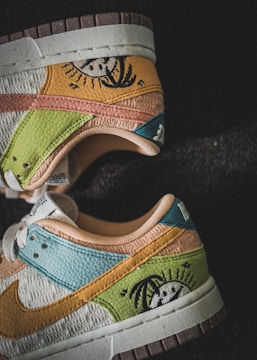 A pair of sneakers with a unique design featuring various colors and a stitched image of a sun with palm trees. The shoes display a mix of leather and fabric materials in shades of yellow, green, blue, orange, and brown. They have a worn, textured appearance and are set against a dark background.
